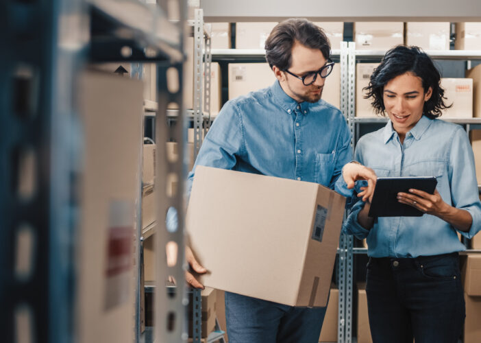 Two retail employees in a warehouse review inventory and logistics on a digital tablet while surrounded by boxes on metal shelves.