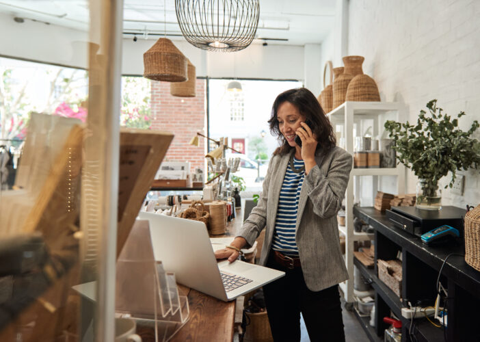 A small business owner in a boutique retail store speaks on the phone while using a laptop at the front counter, surrounded by decor items and retail products.