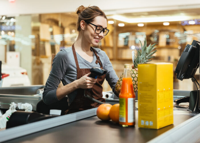 Retail associate scans groceries at checkout, representing secure payment experiences across physical and online retail environments. Payment network security matters.